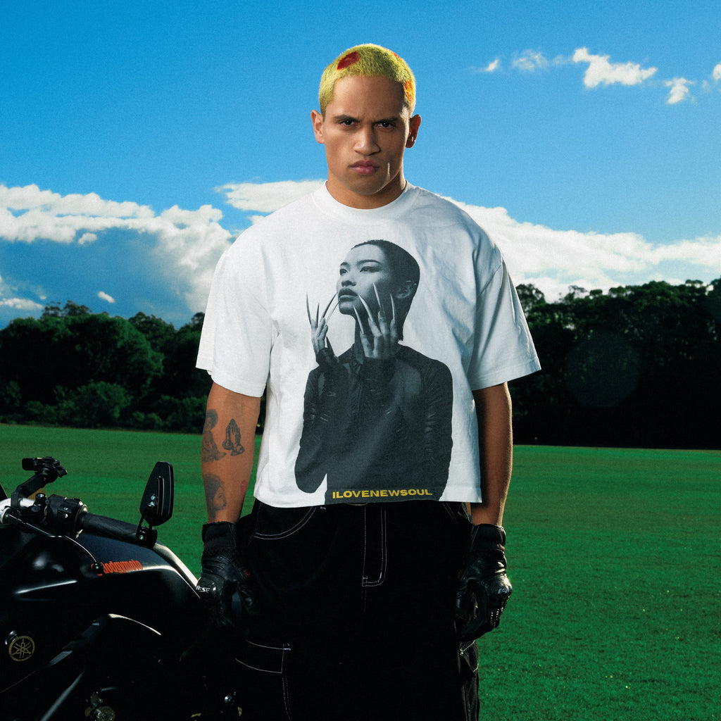 Person wearing a white t-shirt with a graphic design, standing next to a motorcycle on grass with trees and blue sky in the background.