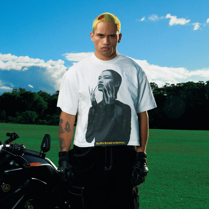 Person wearing a white t-shirt with a graphic design, standing next to a motorcycle on grass with trees and blue sky in the background.