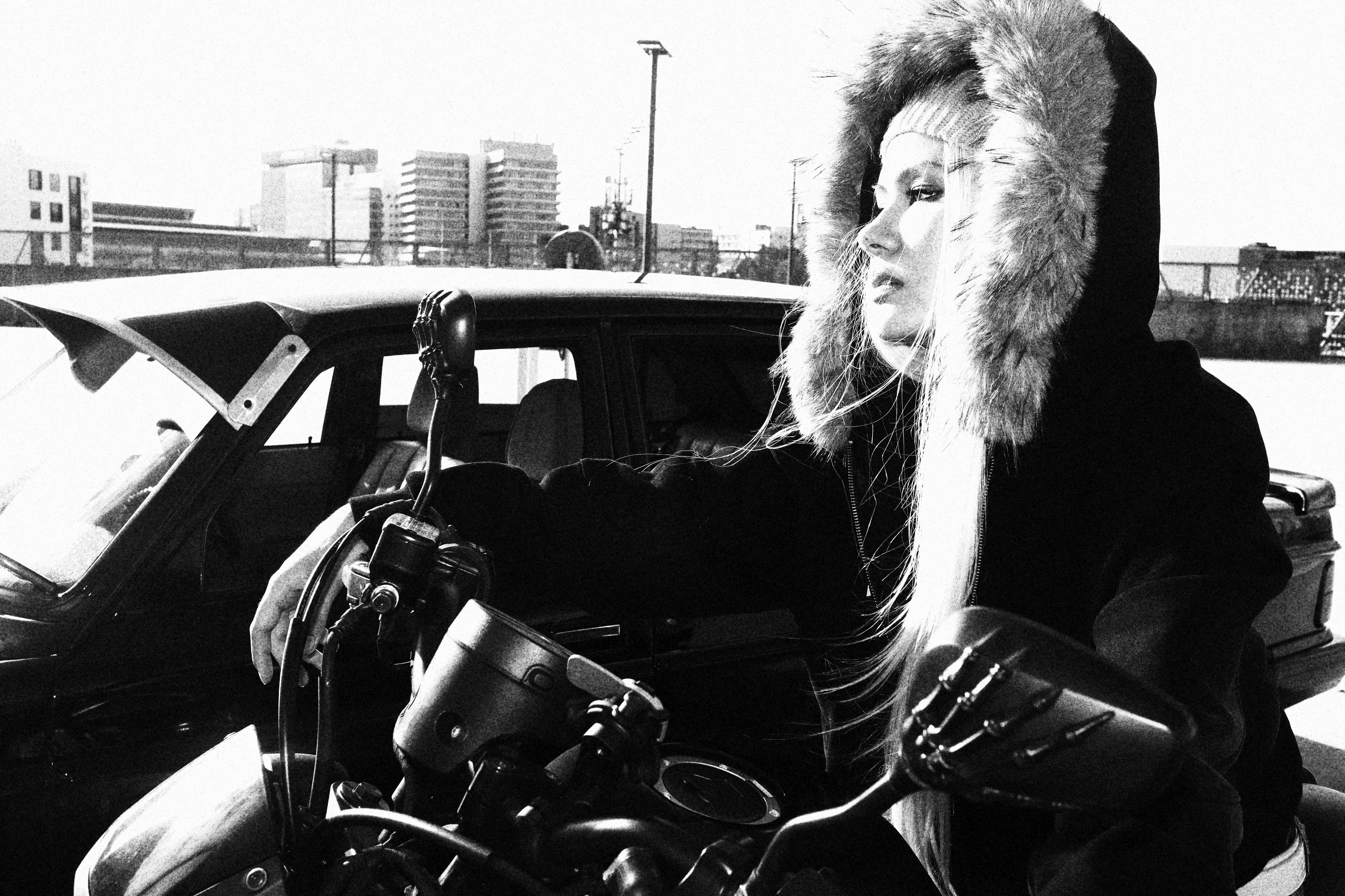 Person wearing a fur-lined coat sitting on a motorcycle with a cityscape in the background